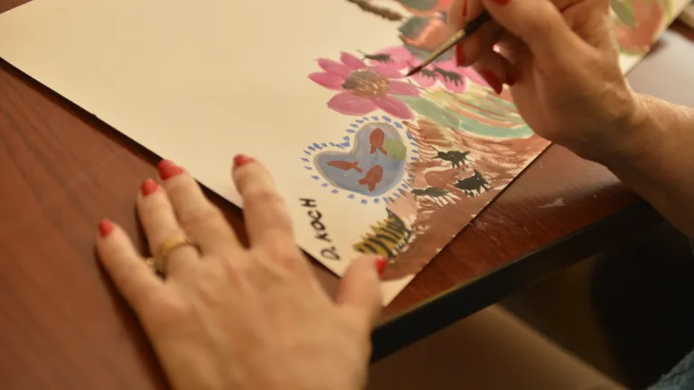 A close up of a womans hands holding a paint brush, a painting of various flowers on a paper in front of them. 