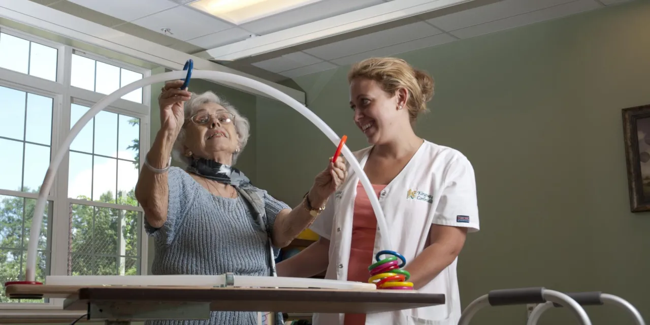 An elderly woman attends a rehab session with a nurse, they are moving colored rings through a curved hoop. 