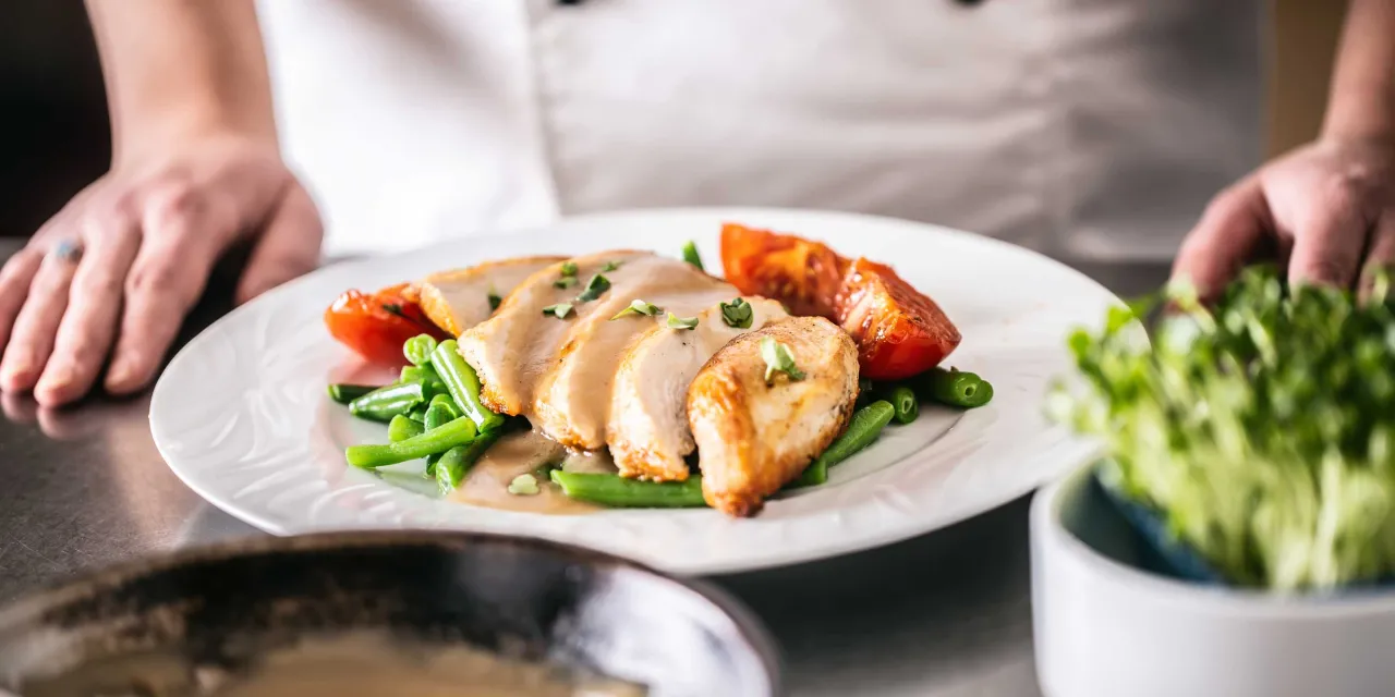 A plate of fine dining food sits on a prep table in front of a chef in a white chefs jacket. 
