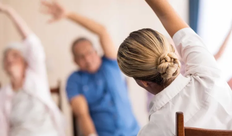 An instructor leads a seated exercise class, two senior residents are seen out of focus in front of her mimicking the move