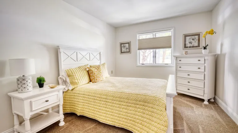 A bright bedroom with beige carpeting, white furniture, and yellow pillows and bedspread