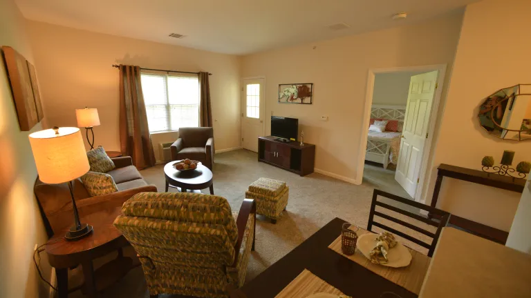 A Kingsway Village living room with brown and green upholstered furniture, a small dining table, and brown drapes