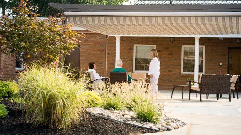 A nurse visits two residents who are sitting outside on the patio