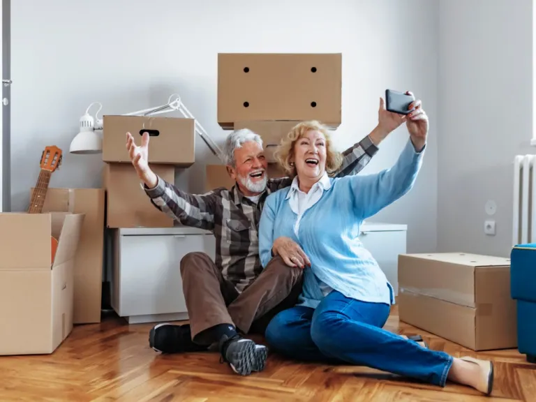 An older couples takes a selfie next to moving boxes