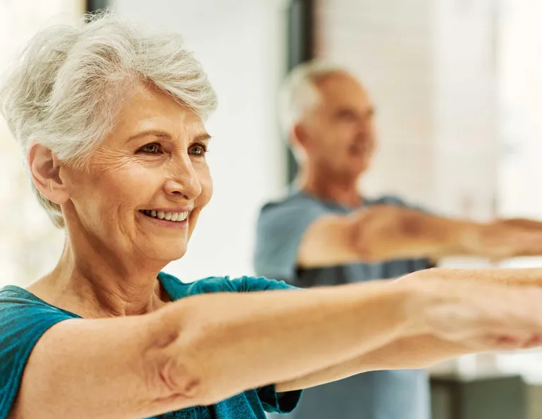 Two seniors enjoy an exercise class, they can be seen smiling with their arms outstretched in front of them. 