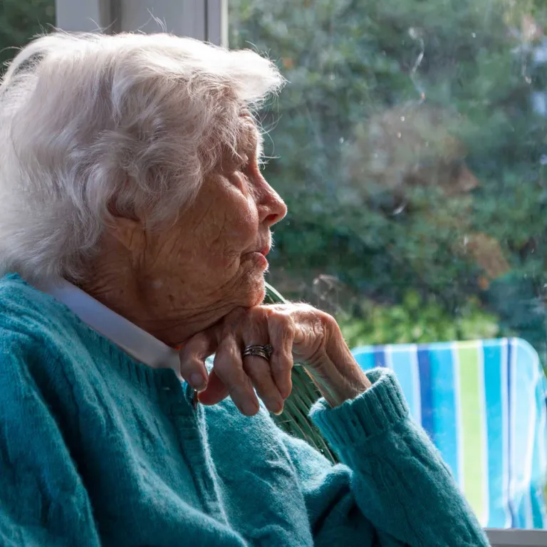 An elderly woman in a blue sweater stares longingly out a window. 