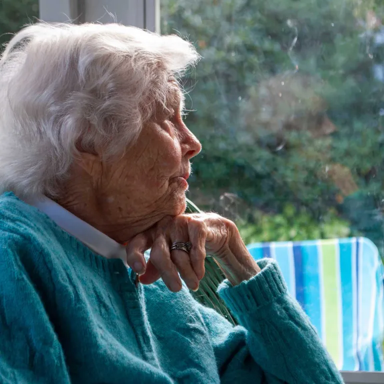 An elderly woman in a blue sweater stares longingly out a window.