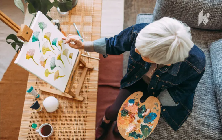 An overhead photo of a senior woman with a paint palette in one hand, a paint brush in the other, working on painting multicolored leaves on a canvas in front of her. 