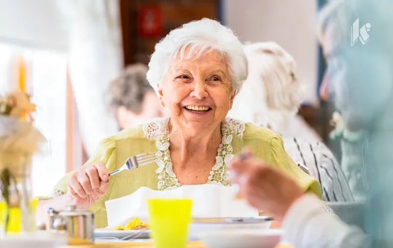 A senior woman sits at a dining table, fork in hand, smiling at a person just behind the camera. 