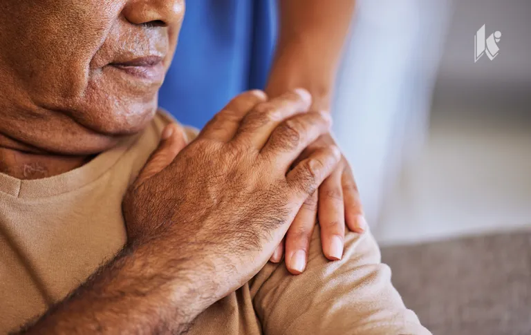 An eldery person can be seen in the foreground, with a nurses hand on their shoulder, their hand is placed atop the nurses hand. 