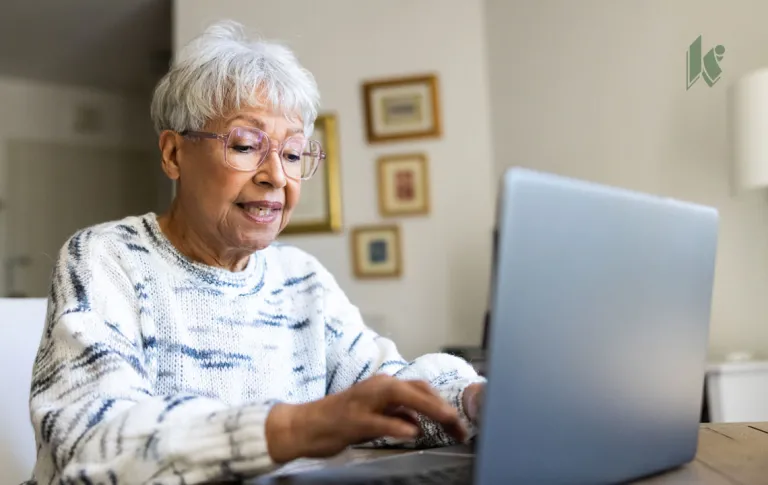 A multiracial senior woman sits in her apartment on a laptop. 