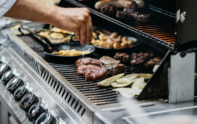 A variety of meats and veggies are on a grill, a hand can be seen hovering above them sprinkling seasoning