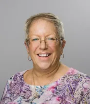 Headshot for Sandra Martin, she has a short haircut and glasses, and is wearing a purple, pink, and blue floral decorated blouse. 