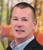 Brian Mott headshot, he is wearing a collared shirt and standing in front of a painting with lots of reds, yellows, and green shades.