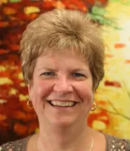 Kathy stands in front of a painting of yellows and reds and greens, smiling at the camera. 