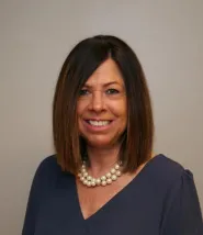 Katie George headshot. Straight brown shoulder length hair, dark blue blouse, pearl necklace. 